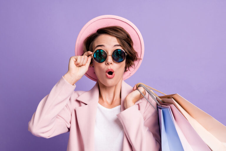Photo of amazed pretty optimistic bob hair young lady hold bags wear glasses pink cap coat isolated on purple color background